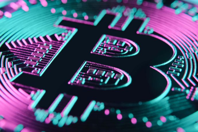 Nahaufnahme einer physischen Bitcoin-Münze, deren ikonisches Bitcoin-Symbol durch neonpinke und blaugrüne Lichteffekte beleuchtet wird.