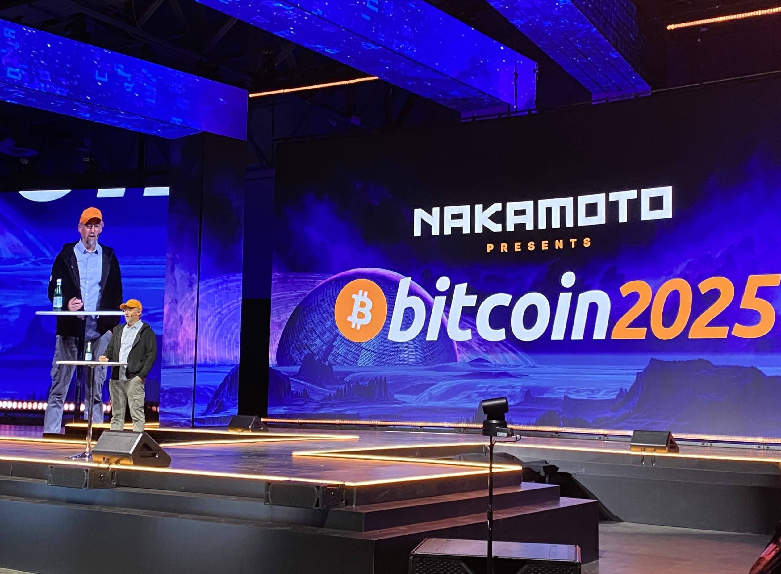 Eine Person mit orangefarbenen Mützen steht auf einer Bühne neben einem großen Bildschirm mit der Aufschrift NAKAMOTO präsentiert Bitcoin 2025 auf einer Konferenz oder einer Veranstaltung zum Thema Bitcoin.