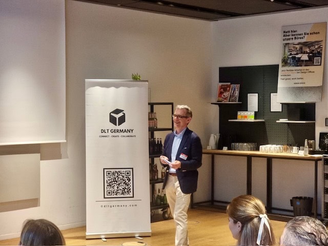 Ein Mann steht neben einem Banner von DLT Deutschland und diskutiert mit einem Publikum über die Zukunft der Kundenbindung in einem modernen Raum mit Regalen, einer Theke, Tassen und einer Kaffeemaschine im Hintergrund.