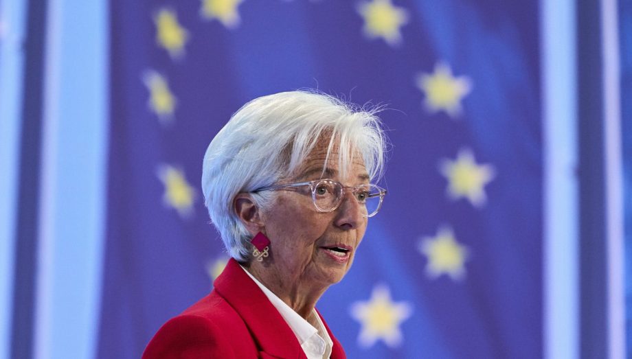 Eine ältere Frau mit kurzen weißen Haaren und Brille, bekleidet mit einem roten Blazer, diskutiert vor einem unscharfen Hintergrund mit gelben Sternen auf blauem Grund über die Inflation der EU-Flagge.