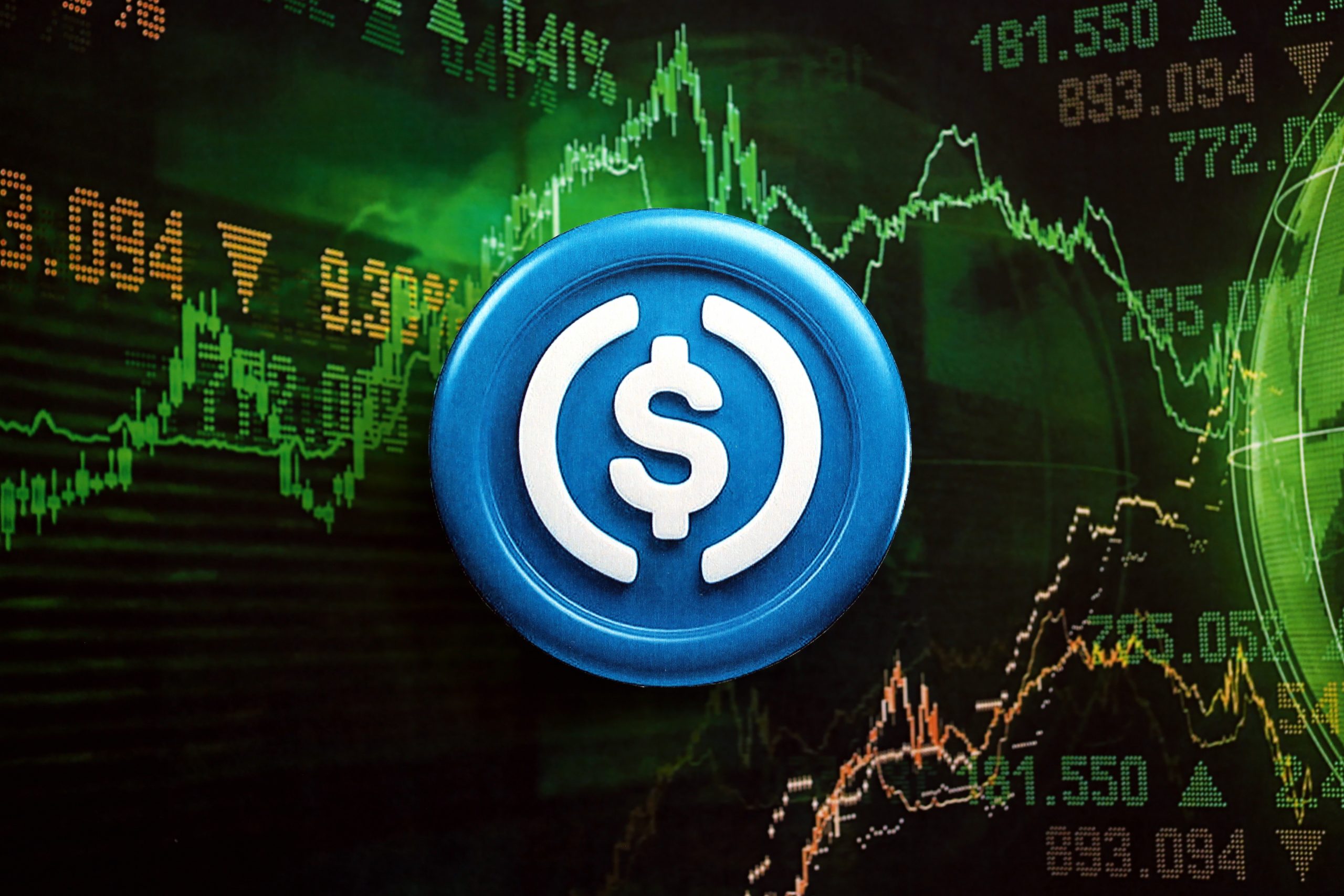 Ein USDC-Münzsymbol, das die beliebte Stablecoin darstellt, wird vor einem digitalen Bildschirm angezeigt, auf dem schwankende Finanz- und Kryptowährungsmarktdiagramme zu sehen sind.