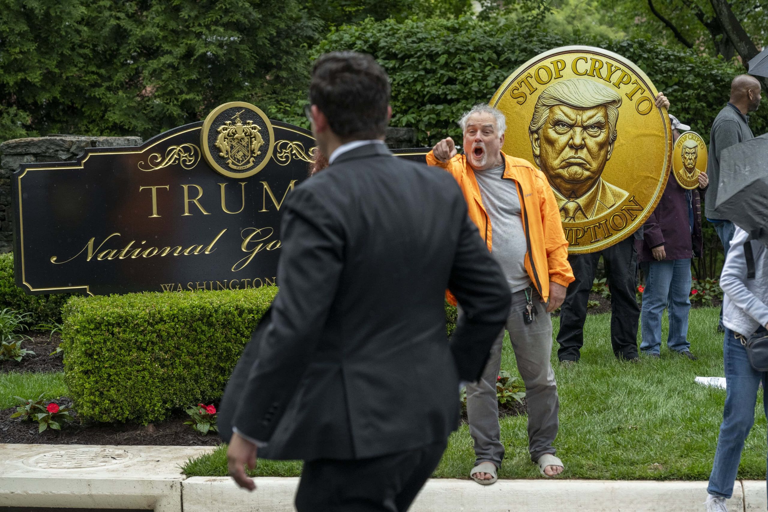 Ein Demonstrant hält eine große Münze mit Donald Trumps Gesicht und der Aufschrift "Stop Crypto Corruption" (Stoppt die Krypto-Korruption), die sich auf das COIN-Gesetz bezieht, vor einem Schild des Trump National Golf Club in die Höhe, während Leute vorbeilaufen.