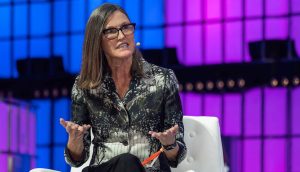 Eine Frau mit langen braunen Haaren und Brille spricht auf einem weißen Stuhl sitzend und gestikuliert mit ihren Händen vor einem Hintergrund aus blauen und violetten Lichtern bei einer Veranstaltung von Coinbase.