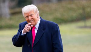 Ein Mann in einem dunklen Mantel und einer leuchtend rosa Krawatte steht draußen auf einer Wiese, lächelt und zeigt in einer für Trump typischen Geste in die Kamera.