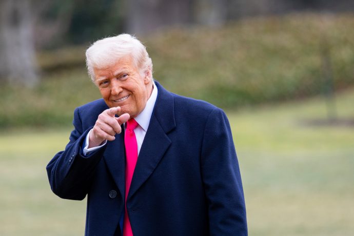 Ein Mann in einem dunklen Mantel und einer leuchtend rosa Krawatte steht draußen auf einer Wiese, lächelt und zeigt in einer für Trump typischen Geste in die Kamera.