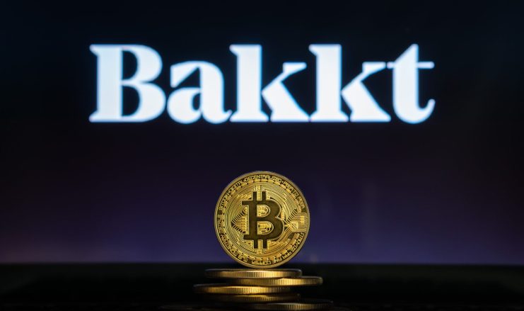 Ein Stapel goldfarbener Bitcoin-Münzen liegt vor einem unscharfen Bildschirm, auf dem das Bakkt-Logo in weißer Schrift zu sehen ist und der die Verbindung zwischen Bitcoin und digitalen Anlageplattformen hervorhebt.