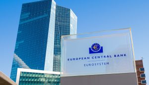 Ein Glasschild mit dem Logo und dem Text der Europäischen Zentralbank (EZB) steht vor einem modernen Bürogebäude aus Glas, das das neue Finanzsystem unter einem strahlend blauen Himmel symbolisiert.