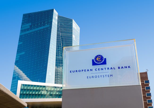 Ein Glasschild mit dem Logo und dem Text der Europäischen Zentralbank (EZB) steht vor einem modernen Bürogebäude aus Glas, das das neue Finanzsystem unter einem strahlend blauen Himmel symbolisiert.