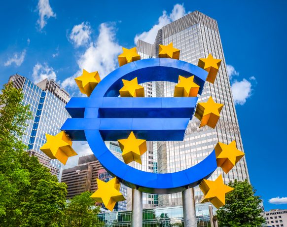 Eine große blaue Euro-Symbol-Skulptur mit gelben Sternen steht vor modernen Bürogebäuden und Bäumen unter einem teilweise bewölkten Himmel und unterstreicht die dynamische Beziehung zwischen traditionellen Währungen und digitalen Assets wie XRP.