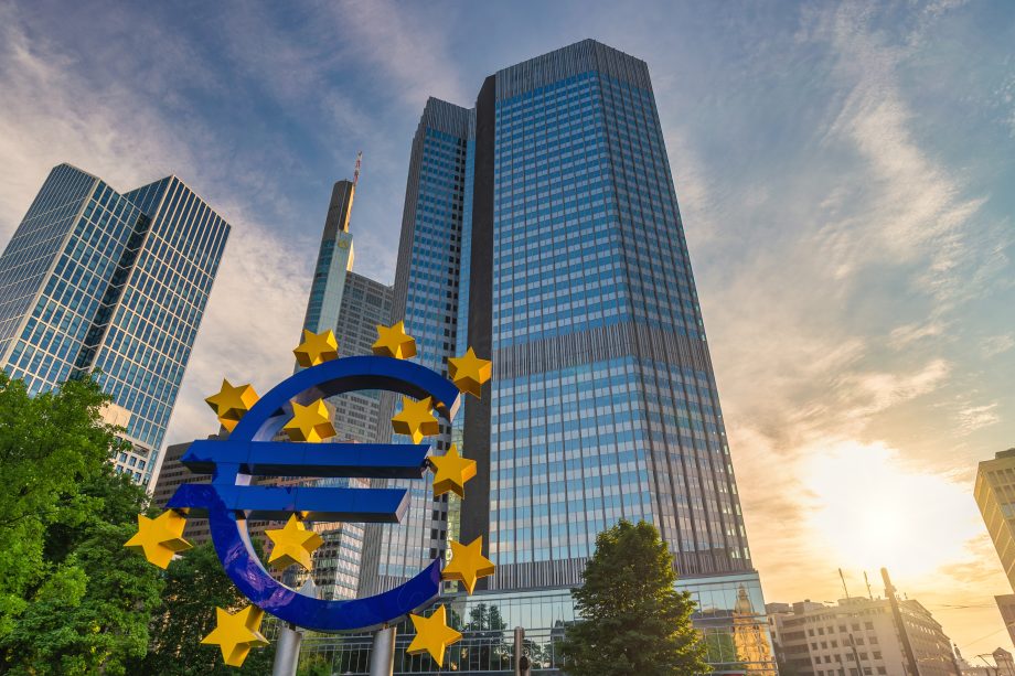 Euro-Symbol-Skulptur vor modernen gläsernen Bürogebäuden, darunter der Morgan Tower, mit der untergehenden Sonne im Hintergrund und einem teilweise bewölkten Himmel.