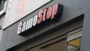Eine Nahaufnahme eines GameStop-Schildes mit der Aufschrift "Game" in weißen Buchstaben und "Stop" in roter Schrift steht über einer teilweise geöffneten Tür, die Kunden in den GameStop-Laden einlädt.