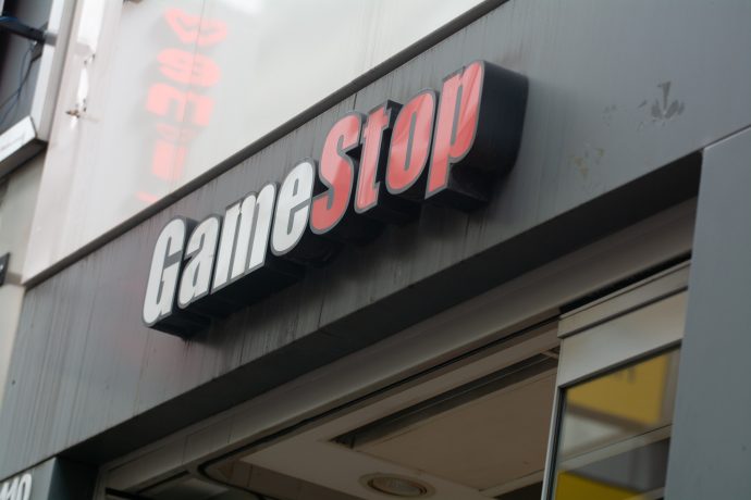 Eine Nahaufnahme eines GameStop-Schildes mit der Aufschrift "Game" in weißen Buchstaben und "Stop" in roter Schrift steht über einer teilweise geöffneten Tür, die Kunden in den GameStop-Laden einlädt.