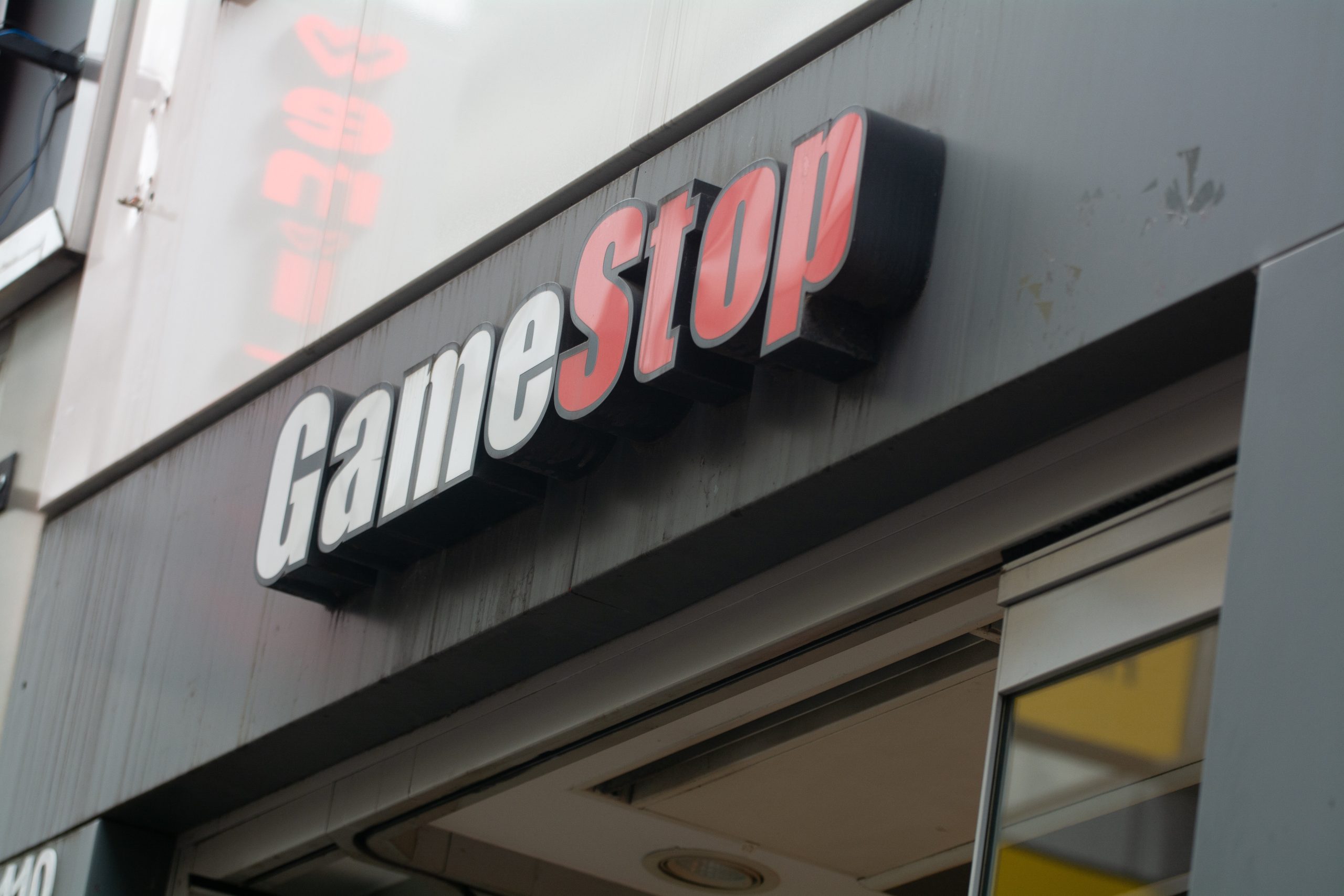 Eine Nahaufnahme eines GameStop-Schildes mit der Aufschrift "Game" in weißen Buchstaben und "Stop" in roter Schrift steht über einer teilweise geöffneten Tür, die Kunden in den GameStop-Laden einlädt.