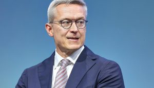 Ein Mann mit kurzen grauen Haaren, Brille und dunkelblauem Anzug mit gestreifter Krawatte blickt vor einem einfarbig blauen Hintergrund zur Seite und verkörpert die Professionalität, die man oft in den Krypto-News DACH-Beiträgen sieht.