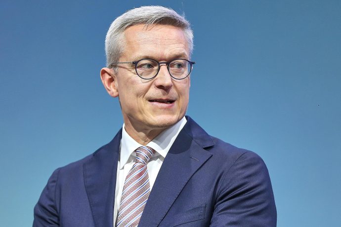 Ein Mann mit kurzen grauen Haaren, Brille und dunkelblauem Anzug mit gestreifter Krawatte blickt vor einem einfarbig blauen Hintergrund zur Seite und verkörpert die Professionalität, die man oft in den Krypto-News DACH-Beiträgen sieht.