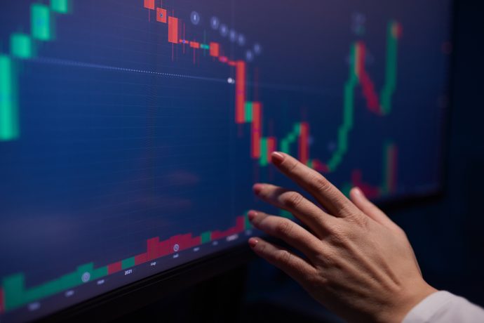 Eine Hand zeigt auf einen digitalen Bildschirm, auf dem ein Candlestick-Diagramm mit roten und grünen Balken zu sehen ist, das Kursbewegungen anzeigt, wie sie beim Krypto-Trading oder am Aktienmarkt üblich sind.