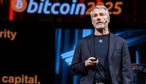 Michael Saylor in einem schwarzen Anzug steht auf der Bühne und hält ein Gerät in der Hand, hinter dem ein auffälliges Bitcoin 2025-Zeichen zu sehen ist.