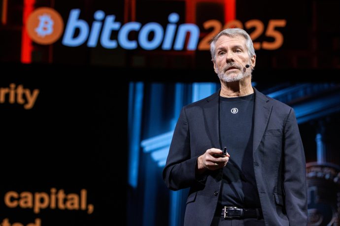 Michael Saylor in einem schwarzen Anzug steht auf der Bühne und hält ein Gerät in der Hand, hinter dem ein auffälliges Bitcoin 2025-Zeichen zu sehen ist.