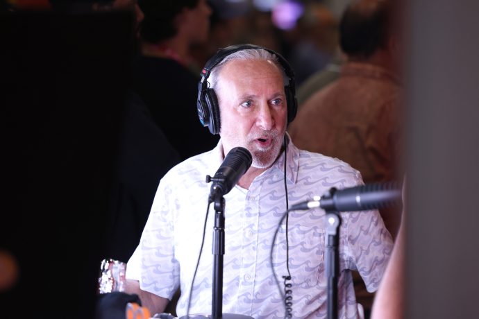 Ein Mann mit Kopfhörern, der Peter Schiff ähnelt, spricht während einer Aufnahme oder eines Interviews in ein Mikrofon und trägt ein gemustertes weißes Hemd.