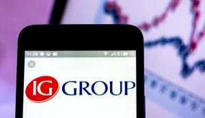 Auf einem Smartphone wird das Logo der IG Group angezeigt, im Hintergrund ist auf einem Computerbildschirm ein verschwommenes Liniendiagramm zu sehen, das möglicherweise Krypto-Trends verfolgt.