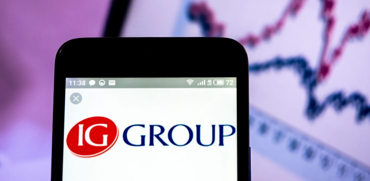 Auf einem Smartphone wird das Logo der IG Group angezeigt, im Hintergrund ist auf einem Computerbildschirm ein verschwommenes Liniendiagramm zu sehen, das möglicherweise Krypto-Trends verfolgt.
