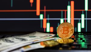 Eine Bitcoin-Münze steht aufrecht neben Dollarscheinen auf einer Tastatur, im Hintergrund wird ein Candlestick-Chart einer Kryptowährung angezeigt.