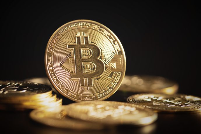 Eine Nahaufnahme mehrerer physischer Bitcoin-Münzen, darunter eine große goldene Bitcoin-Münze, die aufrecht zwischen anderen Münzen auf einer dunklen Oberfläche steht.