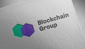 Geprägtes Logo der Blockchain Group mit sich überlappenden grünen und violetten Sechsecken auf strukturiertem weißem Papier, mit einem dezenten, von Bitcoin inspirierten Design.