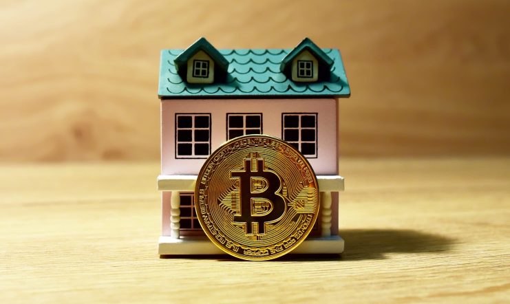 Ein kleines Modellhaus mit einer großen goldenen Bitcoin-Münze, die Krypto-Reichtum symbolisiert, steht aufrecht davor auf einer Holzfläche.