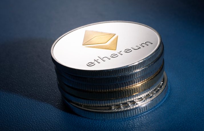 Ein Stapel physischer Münzen mit dem Ethereum-Logo und dem Wort Ethereum, das auf der obersten Münze eingraviert ist, auf einer blauen Oberfläche platziert.