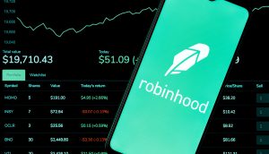 Auf einem Smartphone wird das Logo der Robinhood-App vor einem Computerbildschirm mit Live-Aktienkursen, Portfoliowerten und einem Diagramm steigender Märkte angezeigt, wodurch die Rolle von Robinhood beim modernen Investieren hervorgehoben wird.