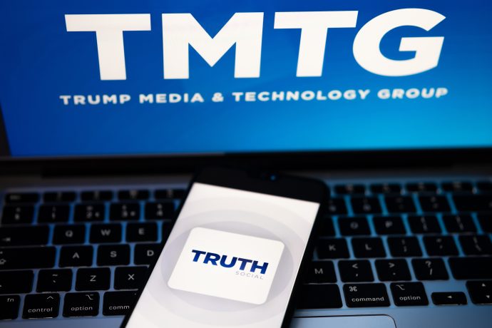 Auf der Tastatur eines Laptops liegt ein Telefon mit dem Logo von Truth Social. Im Hintergrund ist auf einem Bildschirm die Trump Media & Technology Group (TMTG) zu sehen.