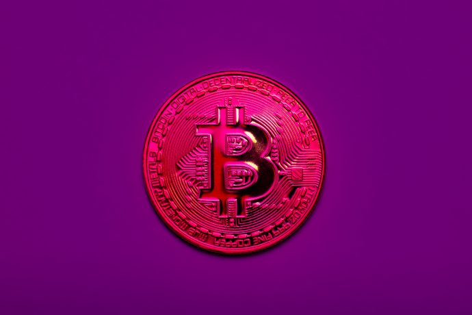 Eine metallisch glänzende Bitcoin-Münze mit geprägten Details wird vor einem einfarbig violetten Hintergrund angezeigt, wodurch das ikonische Währungssymbol hervorgehoben wird.