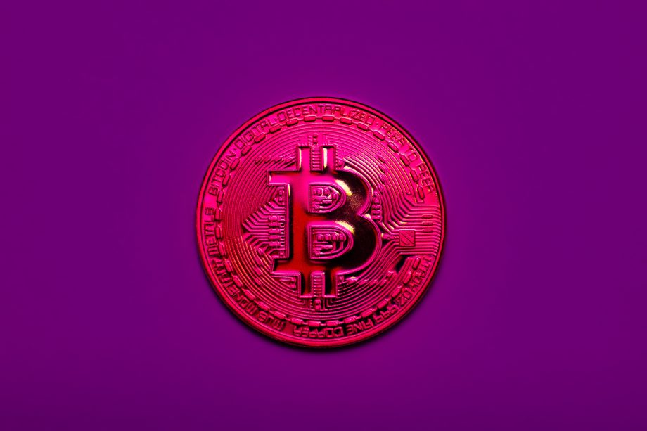 Eine metallisch glänzende Bitcoin-Münze mit geprägten Details wird vor einem einfarbig violetten Hintergrund angezeigt, wodurch das ikonische Währungssymbol hervorgehoben wird.