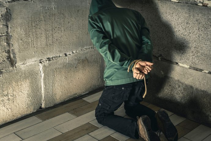 Eine Person in einer grünen Jacke, vermutlich ein Krypto-Influencer, kniet mit auf dem Rücken gefesselten Händen auf dem Boden und blickt in einem schwach beleuchteten Raum auf eine Betonwand – möglicherweise der Schauplatz einer Entführung.