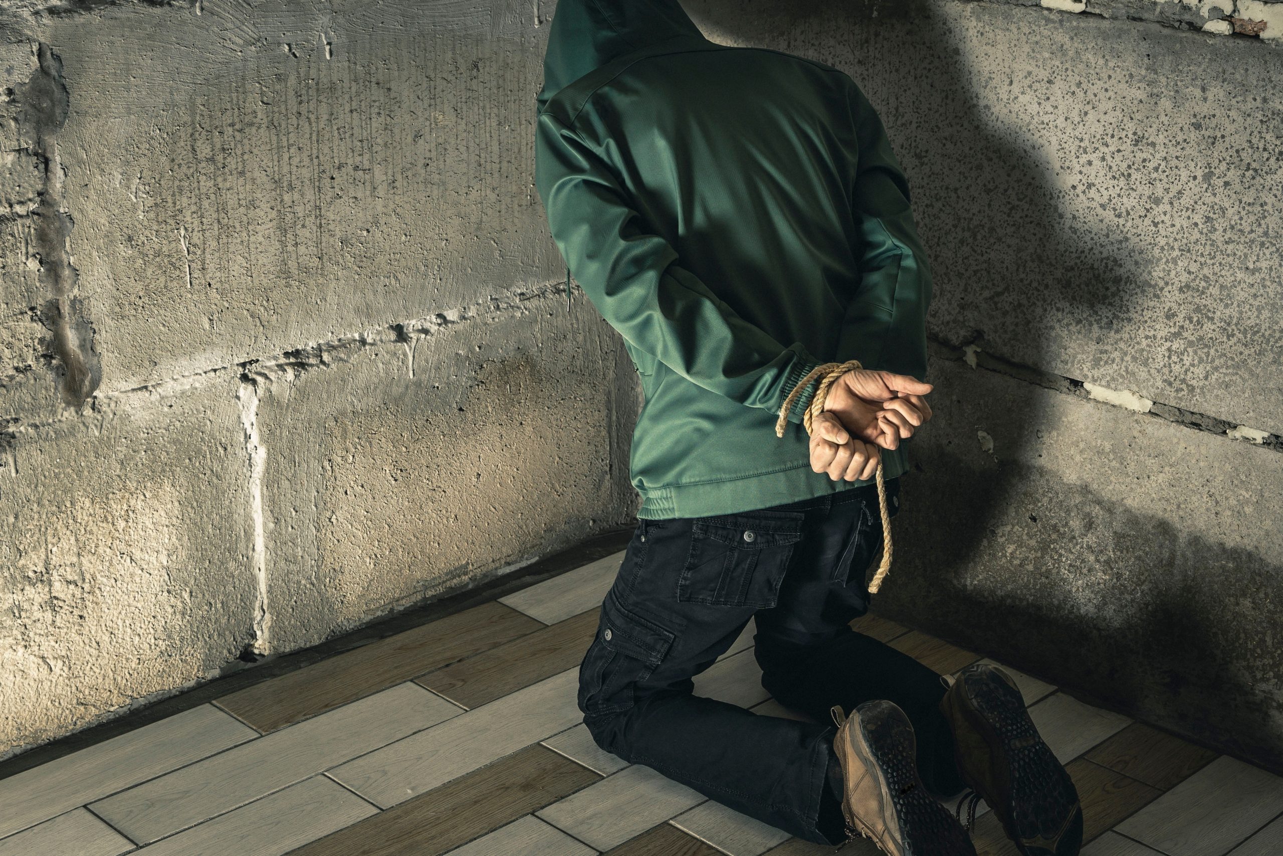 Eine Person in einer grünen Jacke, vermutlich ein Krypto-Influencer, kniet mit auf dem Rücken gefesselten Händen auf dem Boden und blickt in einem schwach beleuchteten Raum auf eine Betonwand – möglicherweise der Schauplatz einer Entführung.
