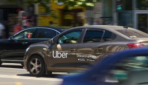 Ein braunes Uber-Auto mit einem „Descargă aplicația“-Schild fährt auf einer Stadtstraße, tagsüber umgeben von anderen Fahrzeugen – modernes Ridesharing, bei dem man sogar mit Stablecoins bezahlen könnte.