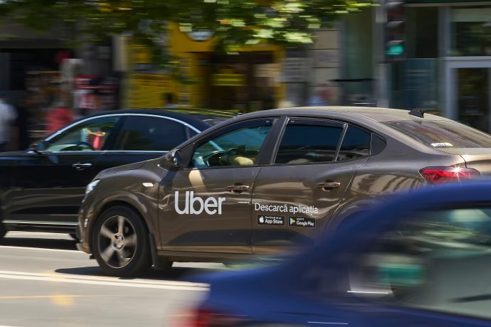 Ein braunes Uber-Auto mit einem „Descargă aplicația“-Schild fährt auf einer Stadtstraße, tagsüber umgeben von anderen Fahrzeugen – modernes Ridesharing, bei dem man sogar mit Stablecoins bezahlen könnte.