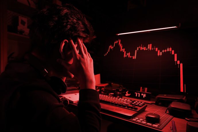 Eine Person sitzt an einem Schreibtisch, blickt auf einen Computerbildschirm in einem schwach beleuchteten Raum und beobachtet den starken Rückgang von Bitcoin auf einem roten Börsenchart.