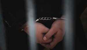 Hinter Metallgittern steht eine Person mit vorn gefesselten Händen und einer dunklen Jacke – vermutlich ein Geiselnehmer.