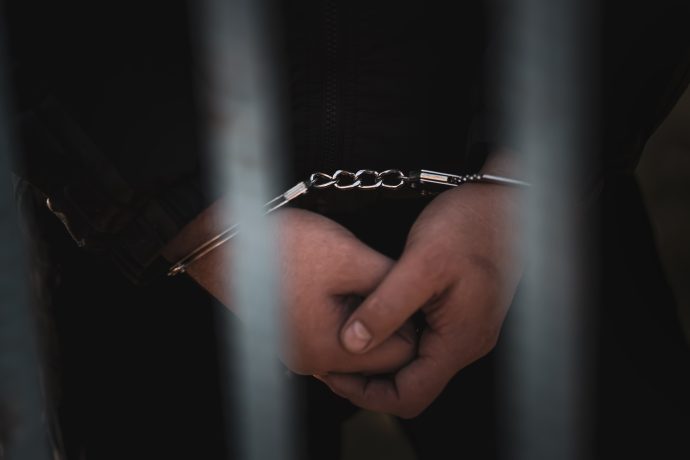 Hinter Metallgittern steht eine Person mit vorn gefesselten Händen und einer dunklen Jacke – vermutlich ein Geiselnehmer.