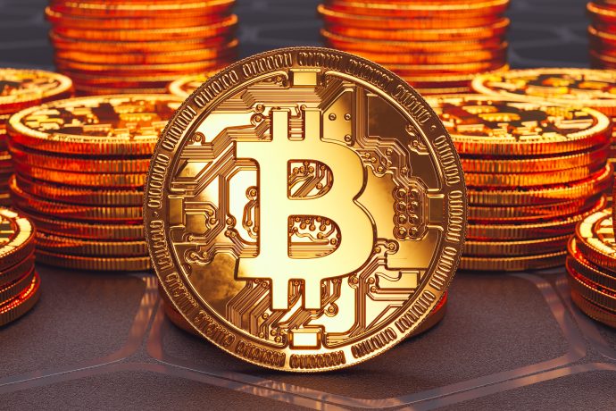 Eine Nahaufnahme einer goldenen Münze mit dem Bitcoin-Symbol, die aufrecht zwischen Stapeln ähnlicher Münzen auf einer dunklen Fläche steht - perfekt zur Illustration einer Bitcoin-Prognose.