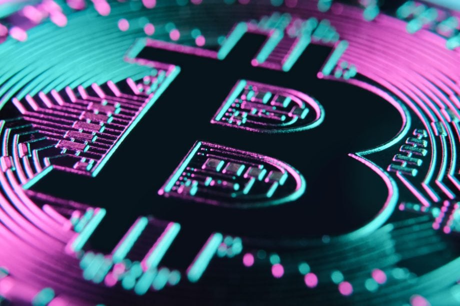 Nahaufnahme eines physischen Bitcoin-Symbols mit teal- und magentafarbenen Lichteffekten, die den Hintergrund des Schaltkreismusters der Münze beleuchten und die futuristische Anziehungskraft von Bitcoin unterstreichen.