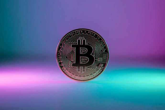 Eine physische Darstellung von Bitcoin steht aufrecht vor einem Hintergrund mit Farbverlauf in Lila und Blaugrün und symbolisiert das Potenzial, in digitale Währung zu investieren.