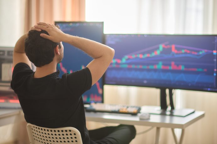 Eine Person sitzt mit den Händen auf dem Kopf an einem Schreibtisch und schaut auf Computermonitore, auf denen Krypto-Trading-Charts und Aktiendaten angezeigt werden. Möglicherweise ist sie gestresst, weil sie einen Denkfehler hat, der ihre Rendite beeinträchtigt.
