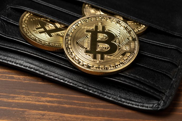 Drei goldfarbene Bitcoin-Token, teilweise in einer schwarzen Lederbrieftasche auf einer Holzoberfläche, ähneln einem automatisch gespeicherten digitalen Vermögenswert in physischer Form.