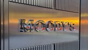Das Foto zeigt eine Metallwand mit MOODYS in reflektierenden Buchstaben, die teilweise einen Sonnenuntergang, eine nahe Straßenszene und Hinweise auf RWA-Aktivitäten in der Umgebung reflektiert.