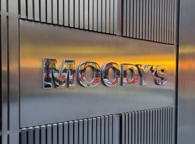 Das Foto zeigt eine Metallwand mit MOODYS in reflektierenden Buchstaben, die teilweise einen Sonnenuntergang, eine nahe Straßenszene und Hinweise auf RWA-Aktivitäten in der Umgebung reflektiert.
