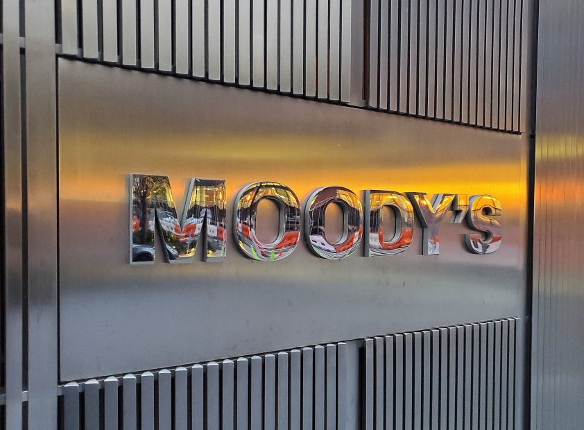 Das Foto zeigt eine Metallwand mit MOODYS in reflektierenden Buchstaben, die teilweise einen Sonnenuntergang, eine nahe Straßenszene und Hinweise auf RWA-Aktivitäten in der Umgebung reflektiert.