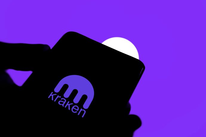 Auf dem Bildschirm eines Smartphones ist das Kraken-Logo zu sehen, das die beliebte Zahlungsapp hervorhebt, und dahinter ist ein lila-weißer Hintergrund zu sehen.